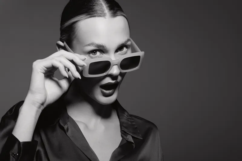 Young white woman with fair skin wearing sunglasses and dark shirt looks surprised in black and white studio portrait. Concept of fashion and expression.