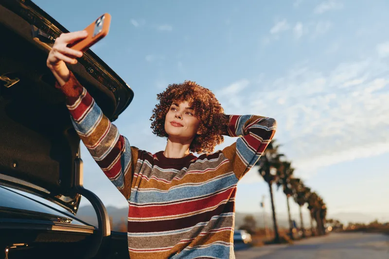 Woman lifestyle portrait, casual young woman with curly hair leaning on a car by street, holding bag strap, travel mood, sunset light over road and palm trees, on quiet vacation afternoon.