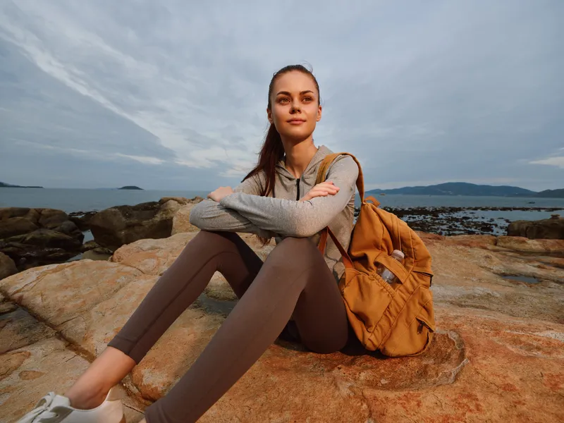 Young woman sitting on rocks by the coast, wearing casual outdoor clothing and smiling, perfect for adventure or lifestyle concepts