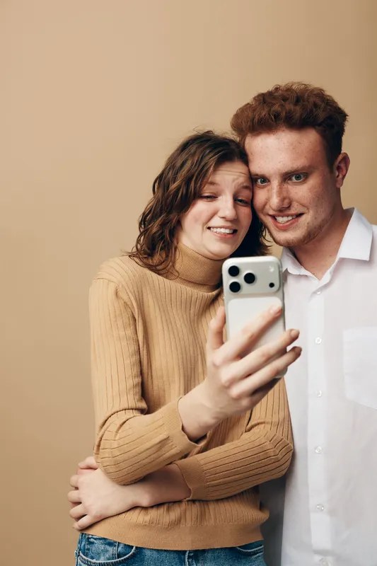 Young couple with curly hair takes a selfie together using a smartphone. They smile and embrace in casual clothes against a plain beige background. Concept of friendship and technology.