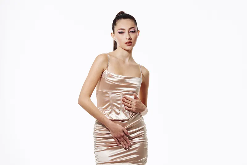 Elegant young woman in a shiny satin dress poses with dramatic makeup and sleek hair on a plain white background. Fashion concept focusing on beauty and creative style.