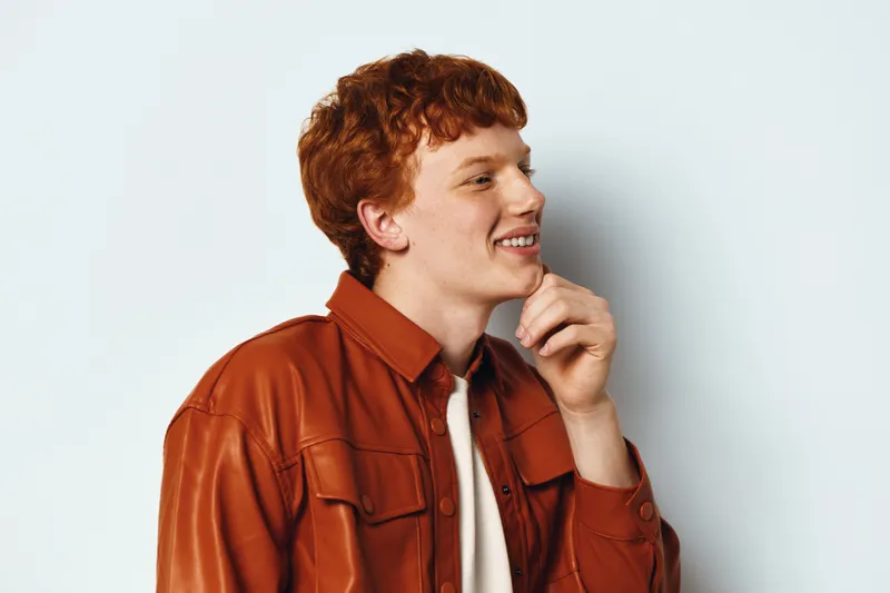 Man with red hair wearing brown leather jacket smiling and touching chin on light background. Casual portrait, side view and thoughtful expression with stylish clothing.