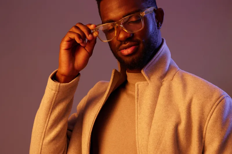 Stylish young Black man wearing glasses and a light coat, expressing confidence against a purple background, perfect for fashion and lifestyle themes