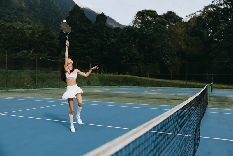 Young female tennis player in a white outfit serving on a blue court, showcasing athleticism and concentration in a vibrant outdoor setting