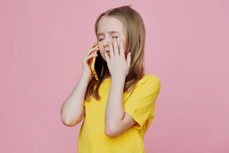 Young girl expressing emotion while talking on the phone, wearing a bright yellow t shirt against a pink background, showcasing joy, connection, and youthfulness