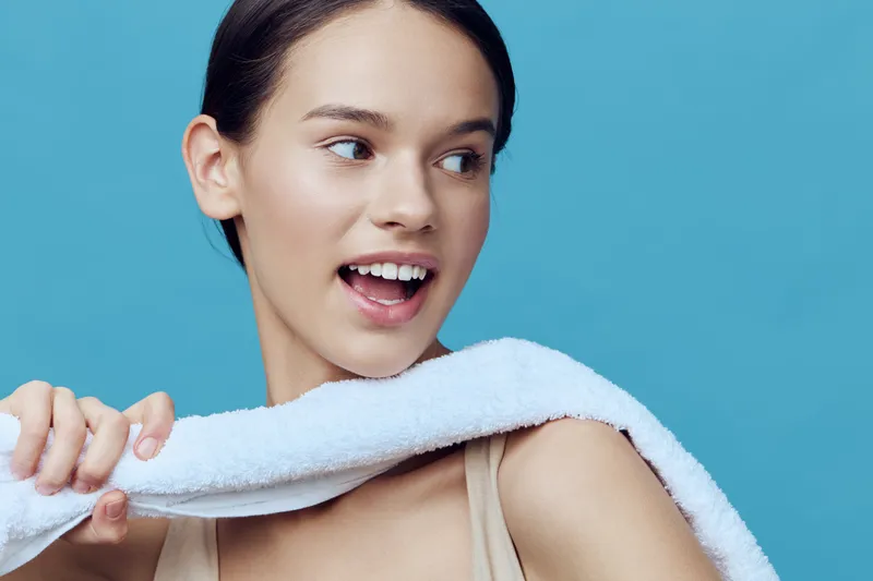 Young woman with a joyful expression holding a towel on her shoulder against a blue background, embodying a fresh and vibrant mood focused on wellness and self care