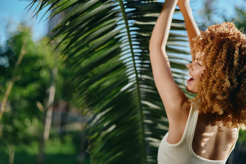 smiling woman with curly hair enjoys tropical nature, sunlight, green palm leaves, and warm weather in outdoor summer setting, expressing happiness and freedom