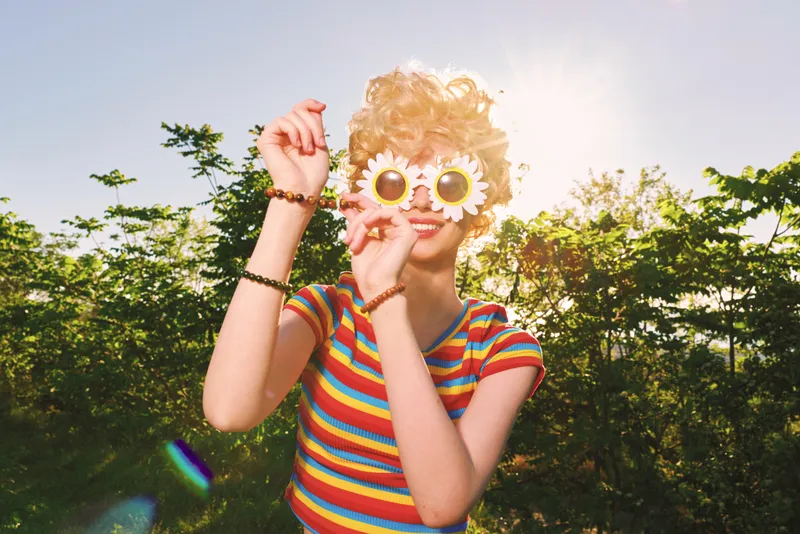 A young woman with blonde curly hair wears round sunflower sunglasses and a colorful striped shirt in bright sunlight, creating a playful retro vibe and a carefree summer mood