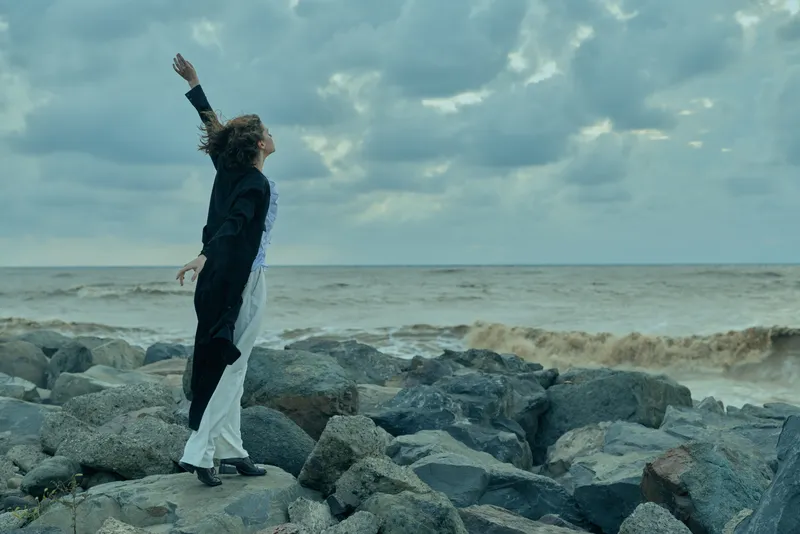 Couple dancing on a rocky shoreline as waves crash and overcast skies loom, a striking contrast between dark suit and flowing white dress, romance by the sea