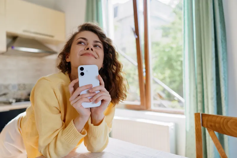 Cheerful woman at home, relaxed indoors, smiling while looking up, warm natural light, cozy sweater, casual pose, sunlit kitchen backdrop, soft colors, lifestyle moment