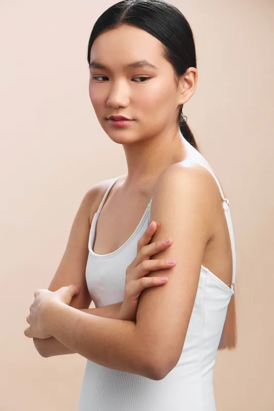 Asian woman with a calm expression stands in a soft studio setting, wearing a light knit top. She gazes downward, conveying quiet confidence and graceful poise.