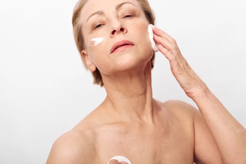 Middle aged woman applying face cream and cleansing skin with cotton pad. Close up on white background. Concept of skincare, beauty routine, self-care, and mature skin health.