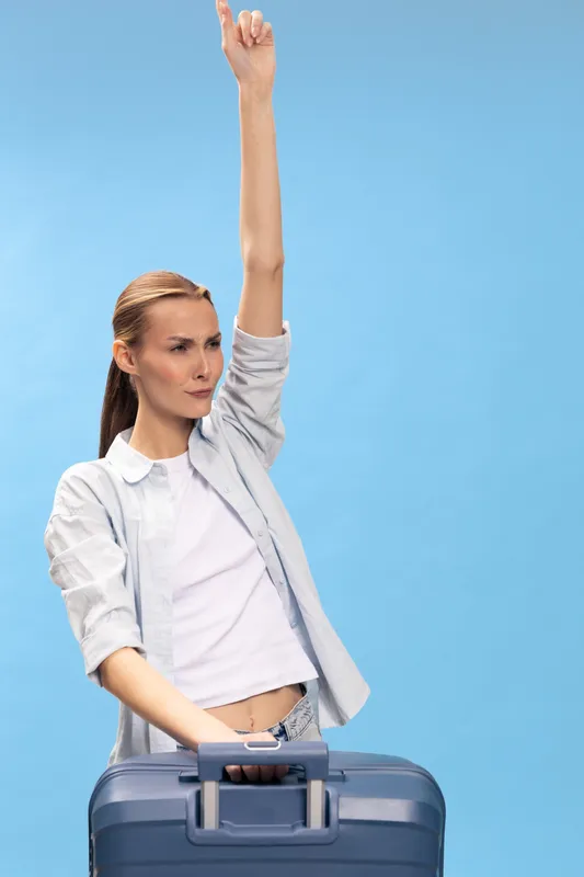 Young woman with long hair holds suitcase handle and raises one arm confidently against blue background. Concept for travel, vacation, freedom, and adventure.