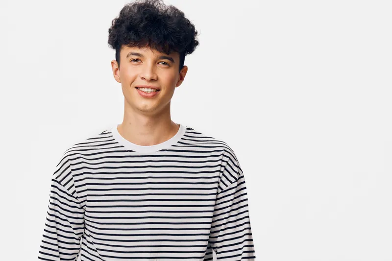 smiling young man with curly hair wearing striped shirt, showing positive emotion and casual style on a white background, natural light and cheerful atmosphere