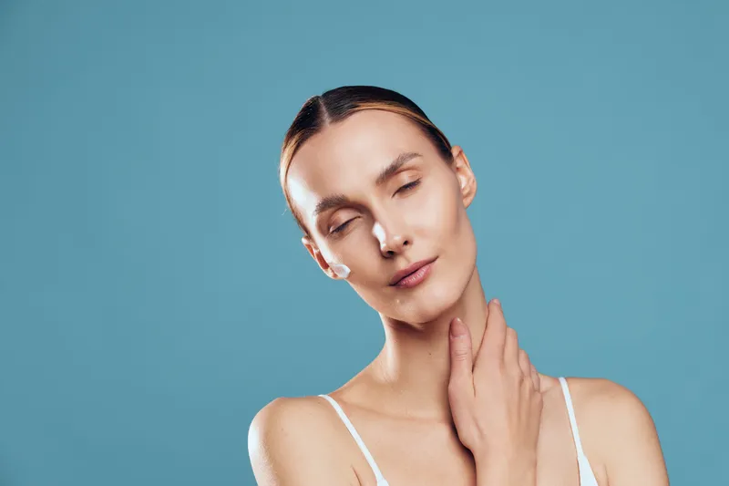 A young woman with clear skin and closed eyes gently touches her neck against a blue background. The image highlights natural beauty, skincare, and relaxation concepts.
