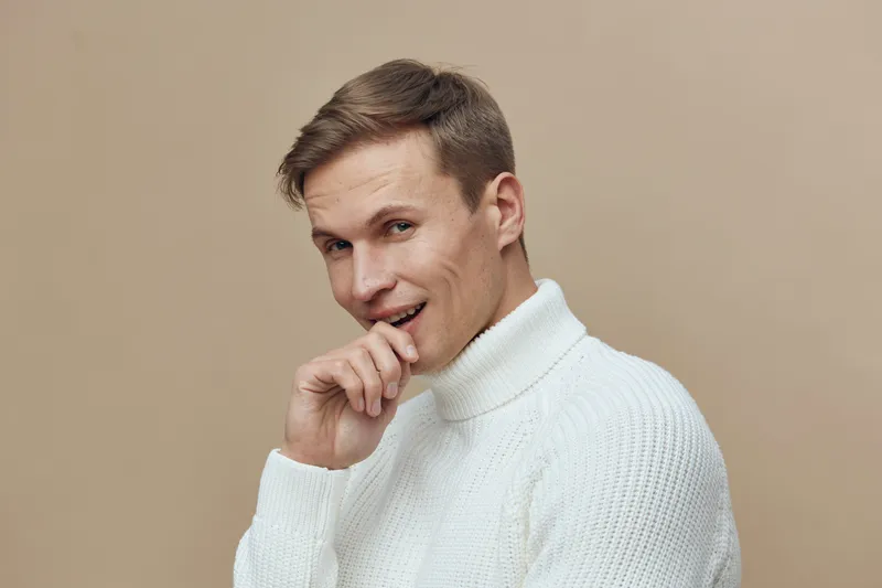 Smiling young Caucasian man wearing white turtleneck sweater looking at camera posing indoors on beige background studio portrait. People lifestyle concept