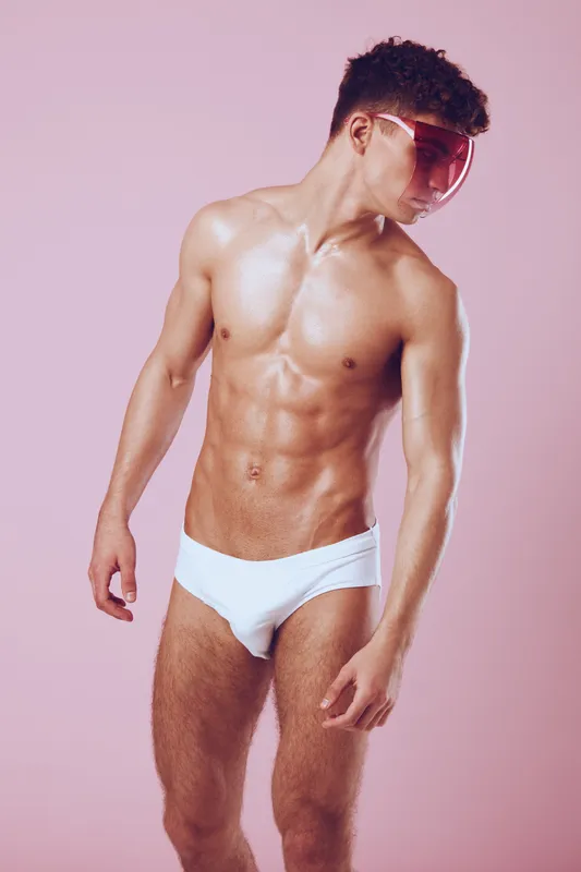 Стильный мужчина-модель в белом плавках и розовых солнцезащитных очках позирует на пастельно-розовом фоне, излучая уверенность и юношескую энергию
