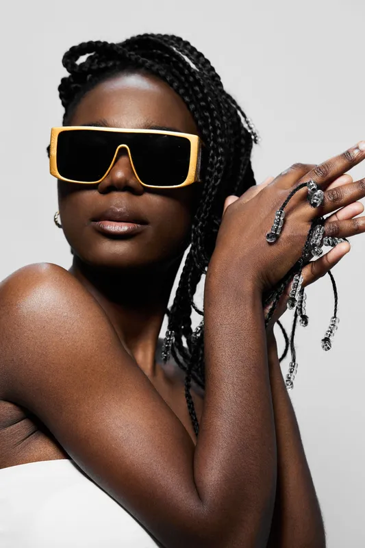 Stylish Black female model with braided hair, wearing oversized yellow sunglasses and demonstrating a confident pose against a minimalist gray background