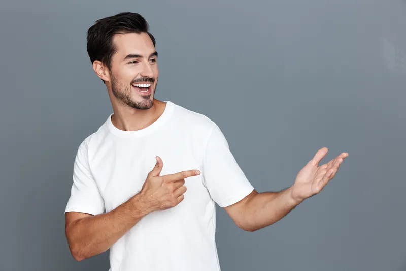 Happy smiling young man in a white t-shirt gesturing with hand while looking at the camera against a plain gray background. People lifestyle concept.