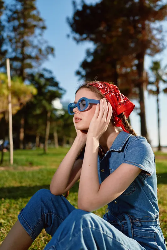 Lifestyle woman in modern boho-western denim street style wearing blue sunglasses and red bandana outdoors with film color effect in a sunny park setting.