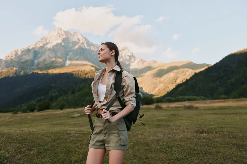 Smiling woman hiking in a meadow with distant mountains, wearing a backpack and casual hiking outfit. Natural outdoor setting conveys adventure, wellness, and exploration inspiring moments