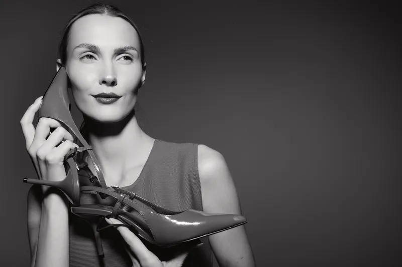 Caucasian woman with light skin holds red high heel shoe near face, looking aside in studio. Concept of fashion, elegance, and style.
