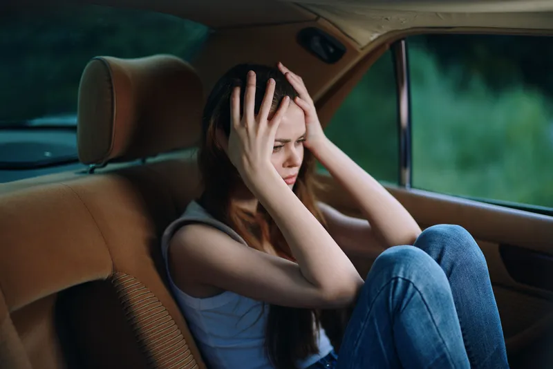 Young woman experiencing stress and anxiety in a car, wearing casual clothing, with a somber expression against a blurred green background, highlighting mental health issues.