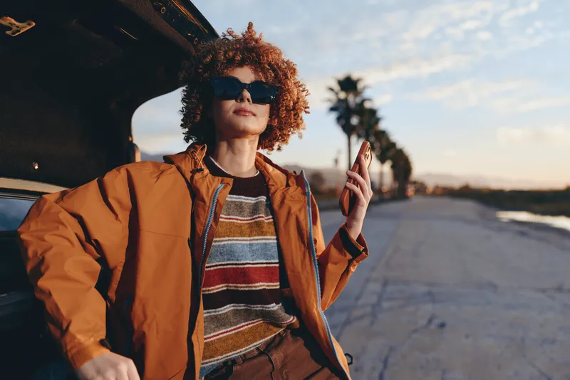 Outdoors lifestyle photo of woman with curly hair wearing sunglasses and orange jacket, posing by road at sunset, casual travel mood, holding car keys, confident smile, cinematic evening light