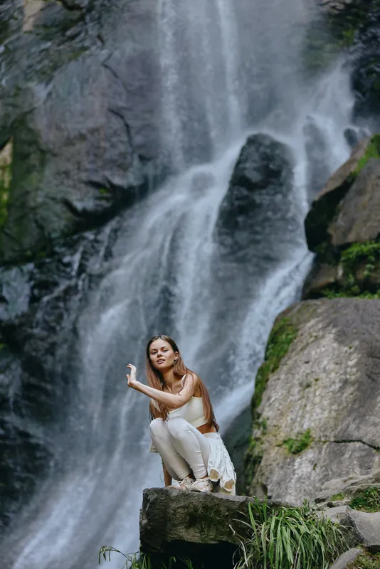 woman near waterfall outdoors wearing white outfit, natural scenery with rocks and flowing water, peaceful and refreshing environment