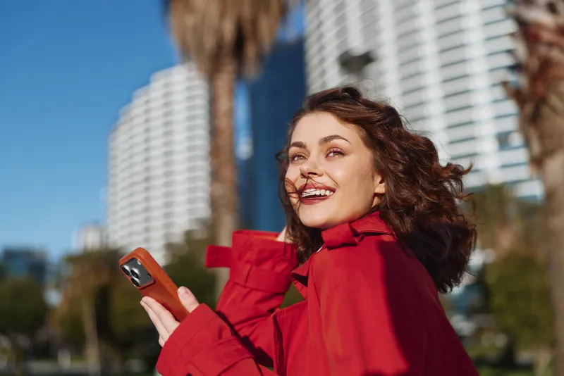 Young woman in red coat smiling outdoors holding smartphone, curly hair blowing in wind, sunny day, modern city background, casual joyful moment
