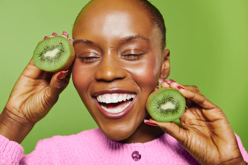 Happy African woman with dark skin holds two halves of kiwi fruit near face, eyes closed and smiling. Bright green background and pink sweater create cheerful mood and healthy lifestyle concept.