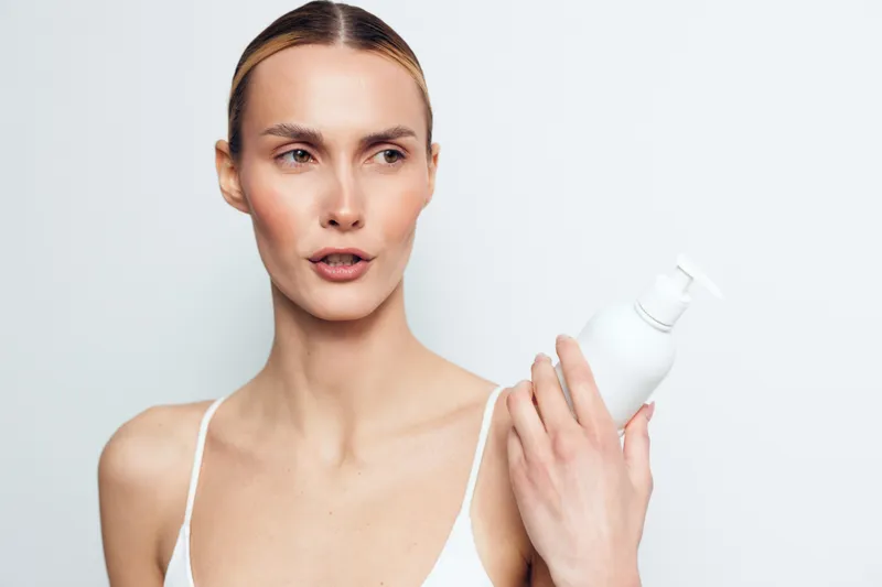A young woman with clear skin holds a white cosmetic bottle while looking to the side against a plain background. Concept for skincare, beauty, and personal care routine.