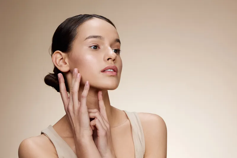 Model demonstrating skincare routine with a natural look, wearing minimal makeup and a beige top, highlighting beauty and self care in a soft, neutral background