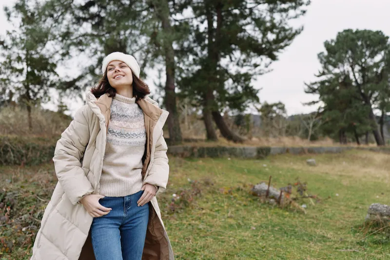 Woman in a cozy coat and knit sweater posing outdoors in a park, with trees and open fields behind her.