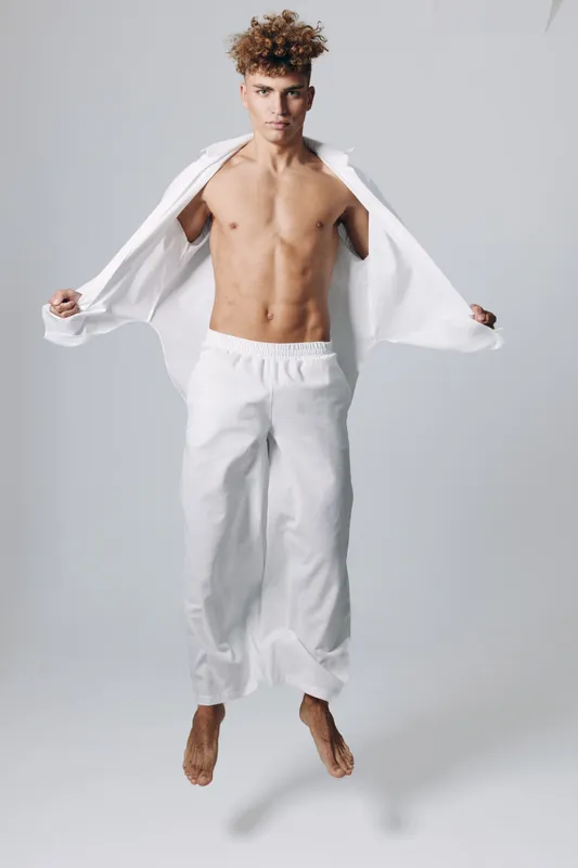 Young man jumping in white loose pants and open shirt with bare chest on gray background, fitness and casual style with curly hair and focused expression in studio.