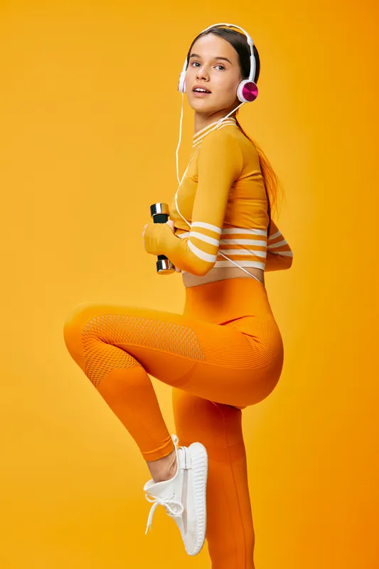 Young woman fitness portrait in vibrant orange activewear holding dumbbells and wearing headphones, single leg pose against seamless yellow studio background with dynamic lighting.