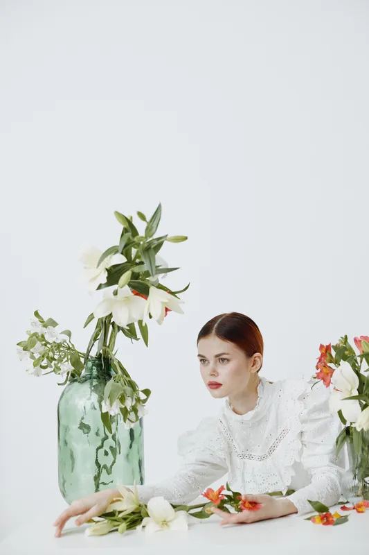 A serene young woman in a white dress arranging vibrant flowers in a glass vase on a minimalistic background, showcasing elegance and floral beauty
