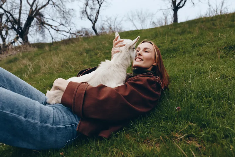 A woman lies on a grassy hillside, smiling as a goat kid nuzzles her, capturing a warm lifestyle moment of affection, companionship, outdoors, nature, friendship, and calm bond.