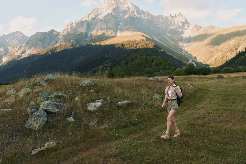 Woman hiking in a mountainous landscape wearing a backpack on a sunny trail. Warm natural lighting highlights rugged terrain, green meadows, and distant peaks. in soft glow.