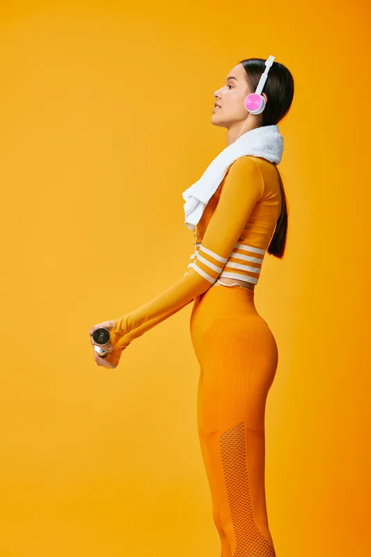 Athletic woman in a bright orange sports outfit with headphones, towel over the shoulder and water bottle in hand, posed mid stride against a vivid orange background.