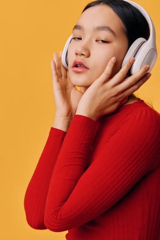 Young woman with headphones enjoying music with closed eyes and hands on ear cups wearing red sweater on orange background. Relaxation, sound, technology, lifestyle and female portrait.