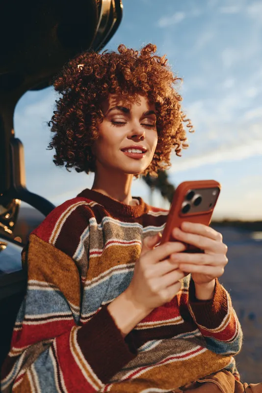 Woman lifestyle portrait outdoors, curly hair in striped sweater, holding smartphone near car at sunset, looking calm and smiling, warm evening light, casual travel moment, relaxation
