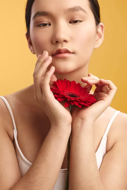 Asian woman holds a red flower near her face in a soft studio portrait with a warm yellow background, conveying calm beauty, focus, and serene mood.