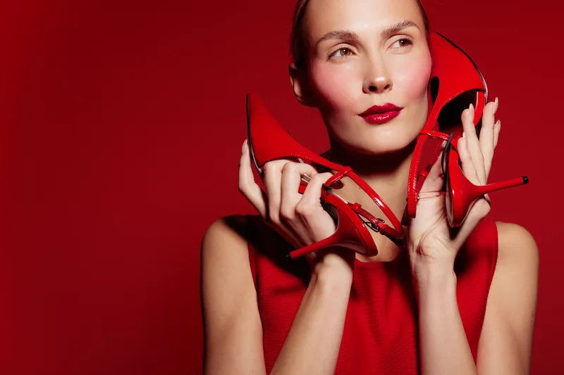 Stylish woman in red dress holds red high heels near face on red background. Fashion concept with bold makeup and elegant accessories in studio lighting.