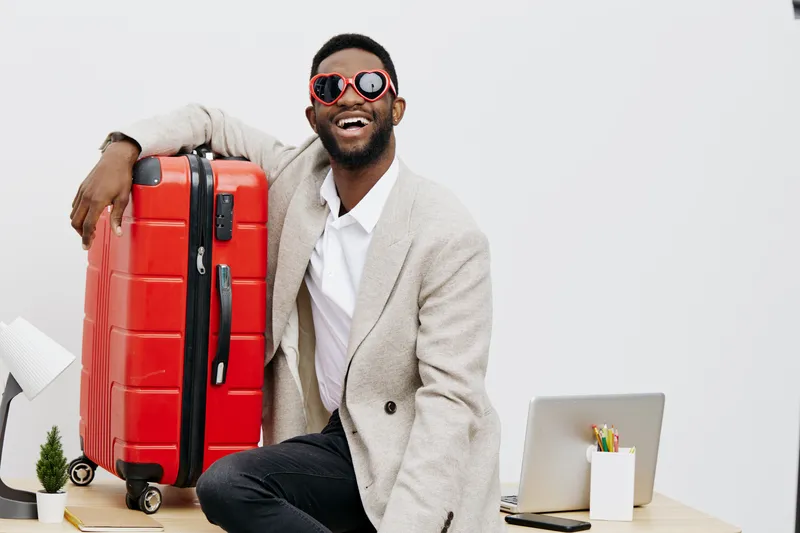 Smiling man with dark hair and beard wearing stylish beige blazer and red sunglasses holding red suitcase in bright minimalist studio setting. Happy traveler ready for adventure, lifestyle, travel con