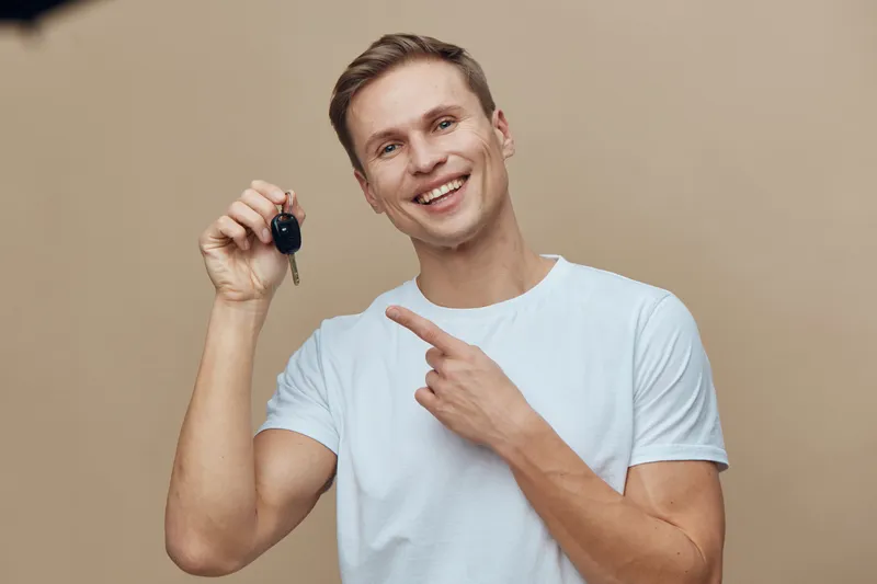 smiling caucasian man hold car key point finger wear white t-shirt isolated beige background digital lifestyle concept