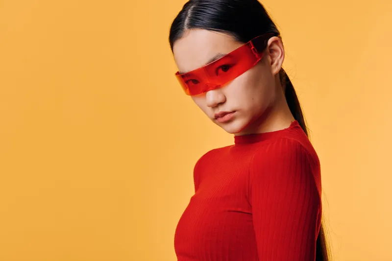 Woman with long dark hair wearing red top and red transparent sunglasses posing on orange background. Stylish young female model looking downward with serious expression and sleek hairstyle.