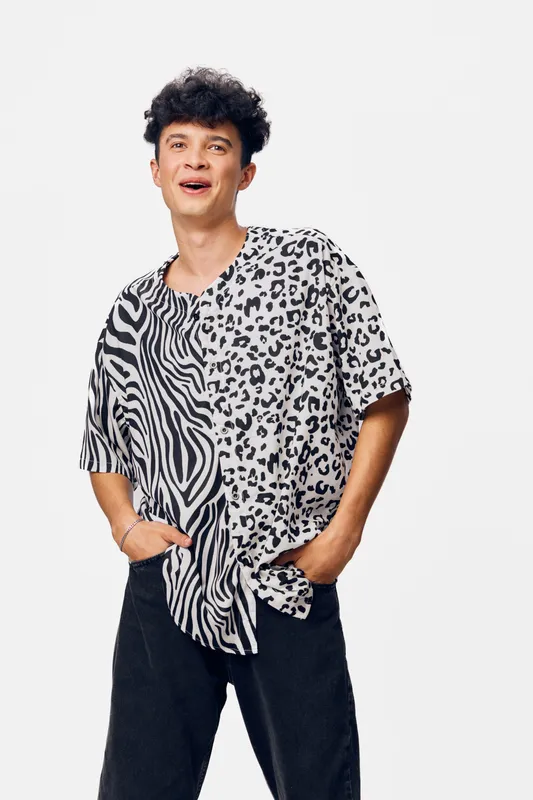 young man wearing a black and white animal print shirt, smiling confidently, posing with hands in pockets against a plain white background