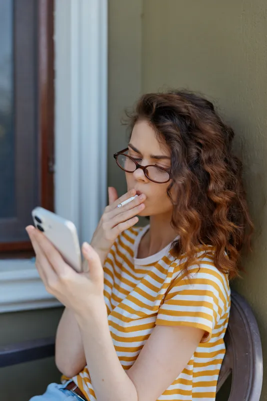 Young woman wearing a striped t shirt sits beside a window, looking at her phone while smoking, capturing a casual candid moment in natural light, relaxed.