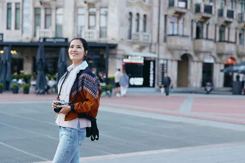 Woman walking on a city street holding a camera, wearing a patterned cardigan and denim jeans. Casual street style captures urban life and a travel mood during daytime.